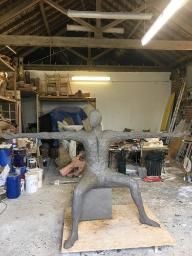 Enlarging Speak to us of Giving in clay - March 2017 Speak to us of Giving in clay. Front view of life size man sitting on cube with elongated arms outstretched. March 2017.