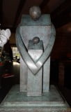 Bronze maquette. Stylised adult standing; arms protectively, round child, both facing us. Green-grey patina. On bronze plinth.