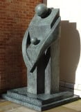 3m high, bronze sculpture. Stylised adult standing, with arms round child, both facing left. Green-grey patina. On plinth.