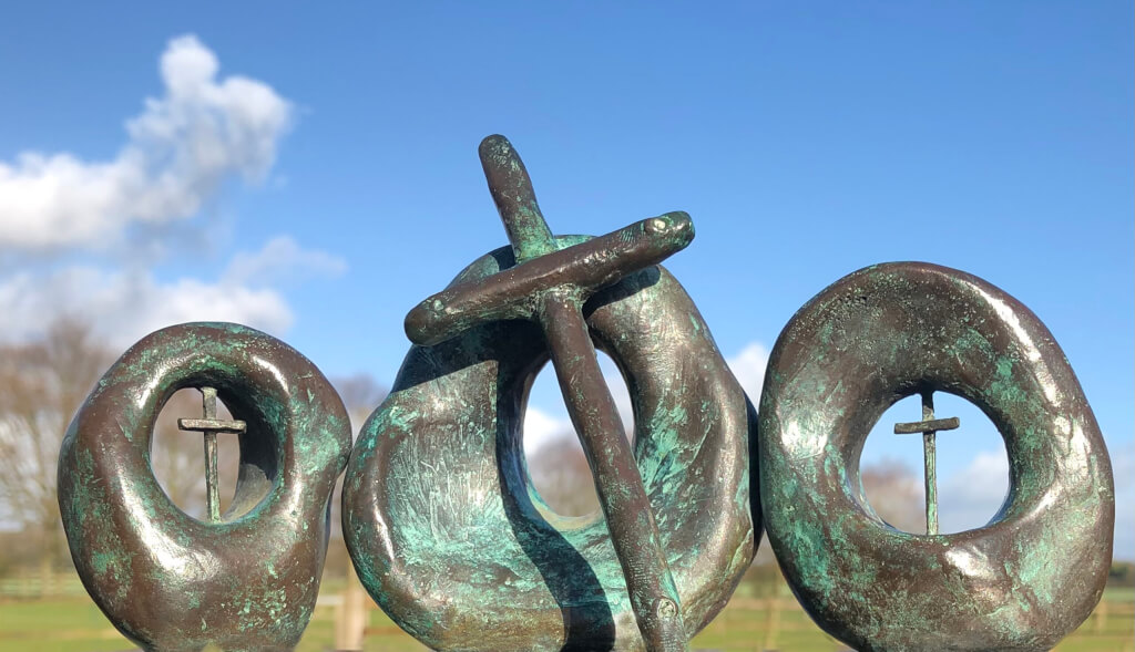 Bronze sculpture showing three circles formed in bronze, the two either side containing small crosses or crucifixes and the central, larger one with a cross taller than the circle, leaning at about 30 degrees to the left or the vertical across the front.